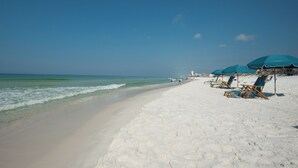 Ein Strand mit weißem Sand, klarem, blauem Wasser und einigen Personen. Entlang des Ufers befinden sich blaue Sonnenschirme und Holzstühle.