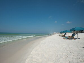 A beach with white sand, clear blue water, and a few people. There are blue umbrellas and wooden chairs along the shore. - 🌴Sandpiper Cove 8246~Steps2beach- 5 Pools~3 Hot Tubs~Tennis Courts~Golf Course⛳ (Destin)