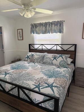 Ein Schlafzimmer mit einem Bett, einem Deckenventilator, einem Fenster mit Jalousien und einem gerahmten Bild an der Wand.