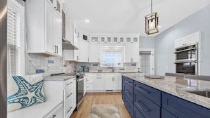 A modern kitchen with white cabinets, a blue accent wall, a large island, and a hanging light fixture. - Mischief Managed NC-  Marsh and Beach views!  Private Pool, Pet Friendly (Oak Island)