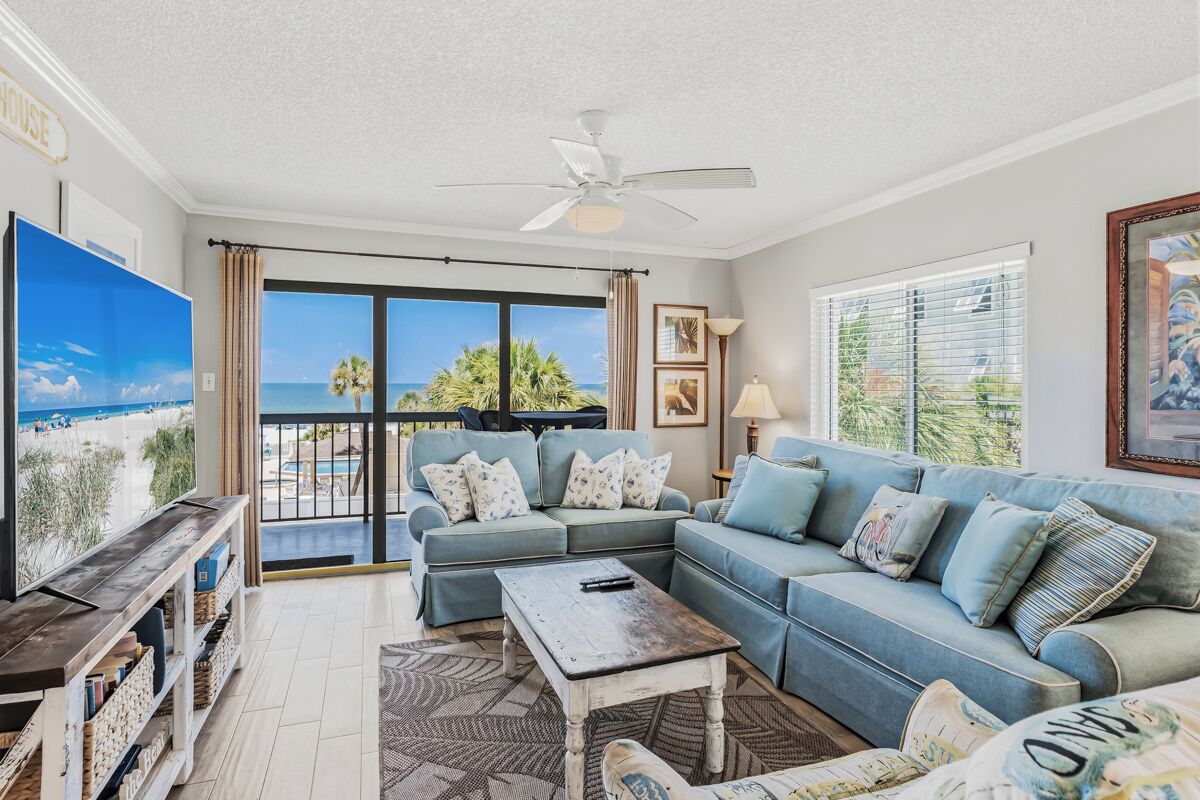 A living room with a large TV, a ceiling fan, a sofa set, and a view of the ocean.