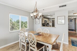 Un coin repas avec une table en bois et des chaises blanches, un lustre et une fenêtre avec des stores.