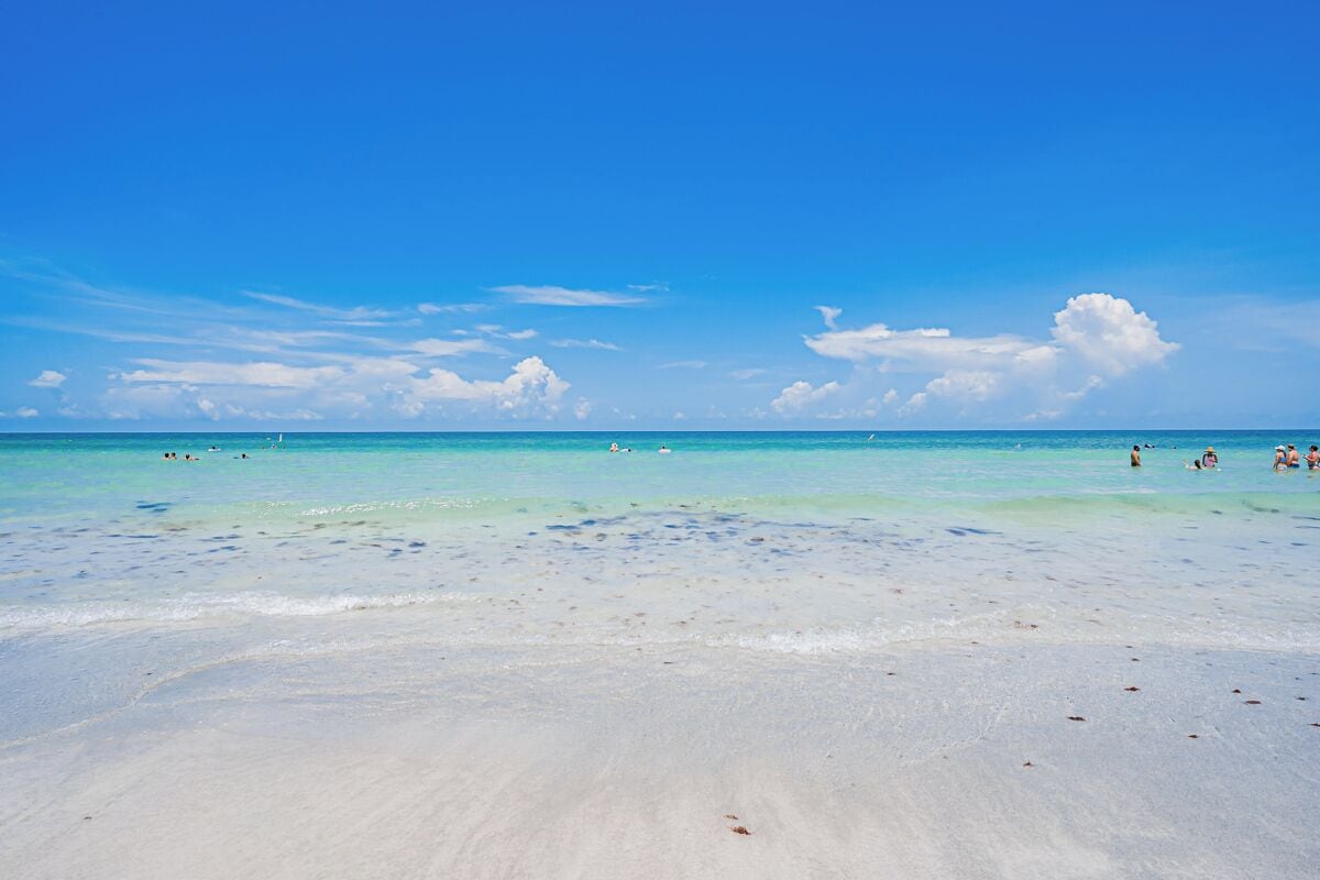 Uma praia com águas turquesa cristalinas, areia branca e algumas pessoas ao longe.