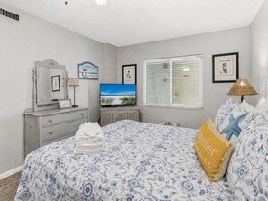 A bedroom with a bed, a dresser, a television, a window with blinds, and a beach-themed sign on the wall. - Gulf Front Condo - Sand Castle North 304 (Indian Rocks Beach)
