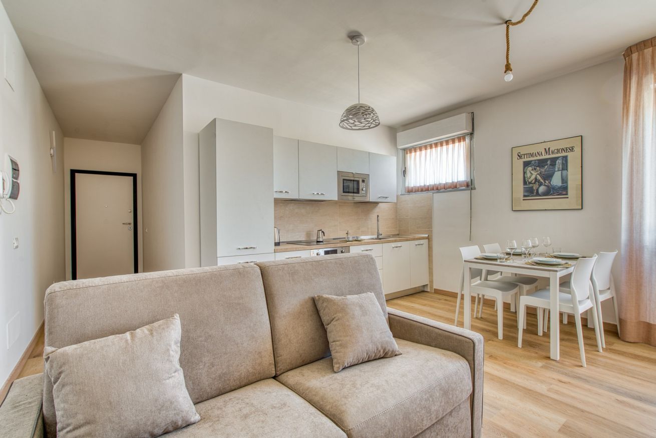 A modern living room with a beige sofa, a dining area with a white table and chairs, and a kitchen with white cabinets and a microwave.