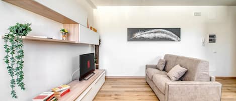 A modern living room with a flat-screen TV, a sofa, wooden flooring, and a wall-mounted shelf with books and a plant.