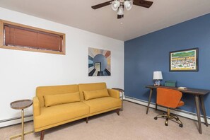 Une pièce avec un canapé jaune, un bureau avec une chaise, un ventilateur de plafond, une fenêtre avec des stores et deux tableaux encadrés accrochés au mur.