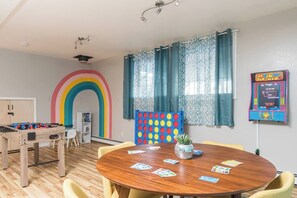 Une pièce avec un baby-foot en bois, une fresque murale aux couleurs de l’arc-en-ciel, un jeu de fléchettes et une borne d’arcade Pac-Man.
