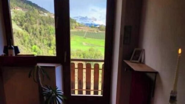 A room with a wooden table, a book, a cup, a candle, and a view of a green landscape through a door.
