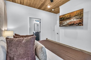 Une chambre d’hôtel comprenant un lit, des oreillers, une table de chevet, une lampe, une salle de bain avec un miroir et un tableau représentant un paysage naturel.