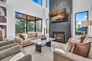A spacious living room with a high ceiling, large windows, a fireplace, and modern furnishings. - Luxury Roof Top Cabin! Splashing Views! Pool! Hot Tub! Gaming Simulator! (Sevierville)