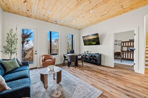 A spacious living room with a wooden ceiling, large windows, a sofa, a dining table, and a TV. - Luxury Roof Top Cabin! Splashing Views! Pool! Hot Tub! Gaming Simulator! (Sevierville)