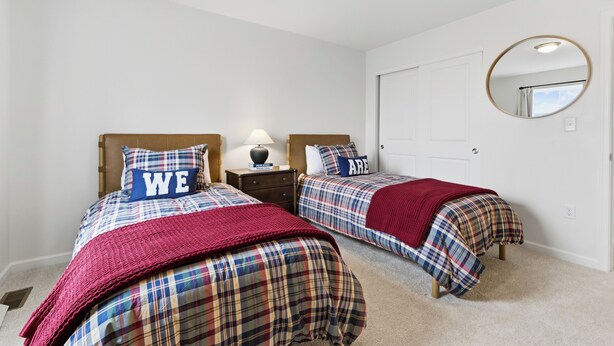 Un dormitorio con dos camas, cada una con edredón y almohadas a cuadros, una mesita de noche con lámpara, y un espejo redondo en la pared.