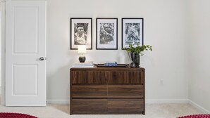 Una habitación con una puerta blanca, una consola de madera con libros y una lámpara, y tres fotografías enmarcadas en la pared.