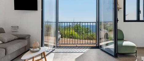 A modern living room with a grey sofa, a small table with a decorative item, a green chair, and a large sliding glass door leading to a balcony with a view of the sea and greenery.