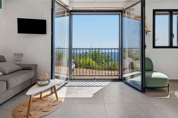 A modern living room with a grey sofa, a small table with a decorative item, a green chair, and a large sliding glass door leading to a balcony with a view of the sea and greenery.