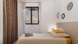 A bedroom with a bed, a window with curtains, a bedside table, and a wall-mounted mirror.