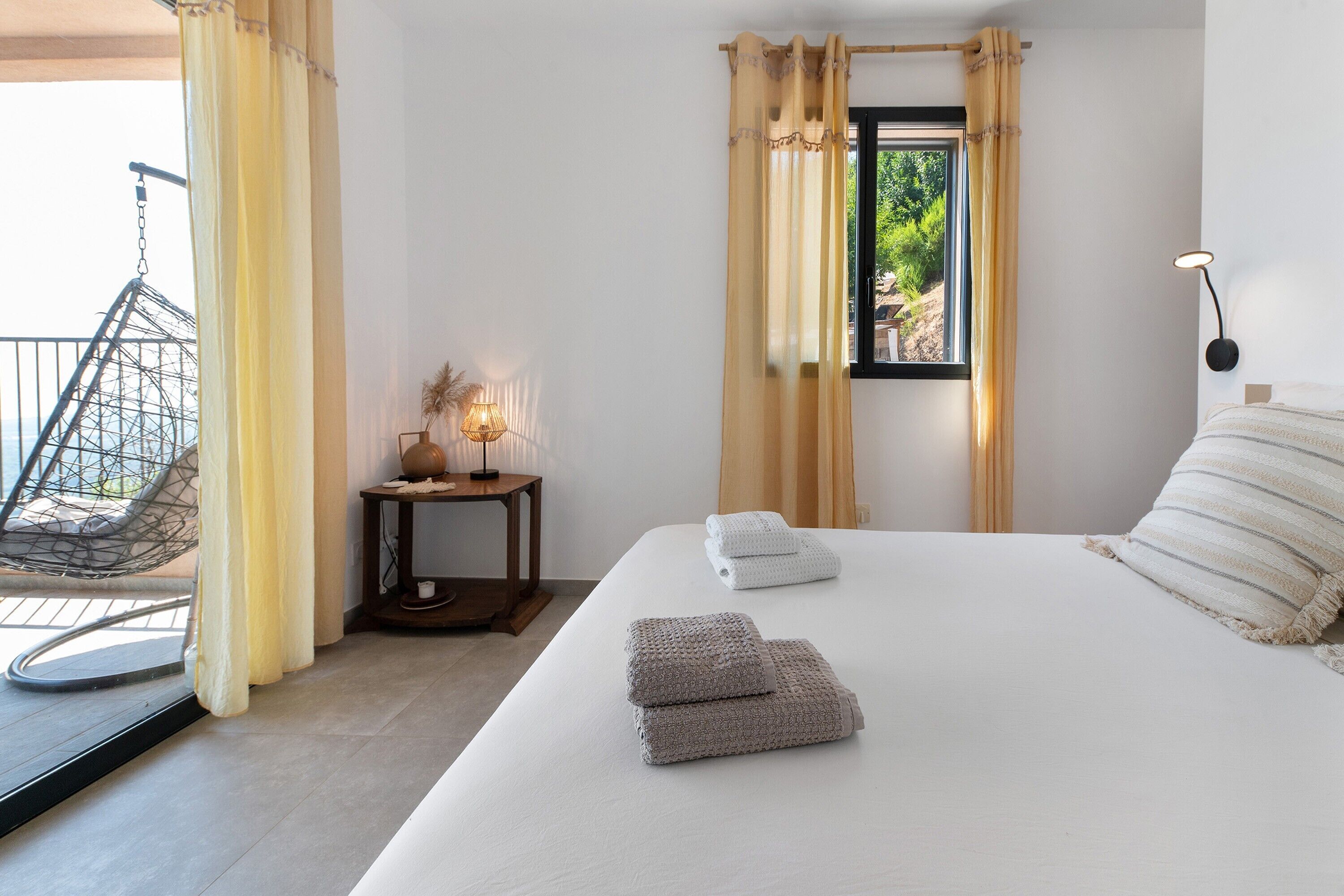 Um quarto com uma cama, duas toalhas, uma mesinha de cabeceira com abajur, uma janela com cortinas e uma cadeira do lado de fora.
