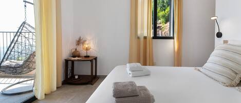 Um quarto com uma cama, duas toalhas, uma mesinha de cabeceira com abajur, uma janela com cortinas e uma cadeira do lado de fora.