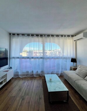 A living room with a TV, a sofa, a coffee table, and a large window with sheer curtains. - Charming studio in marvelous La Grande-Motte with AC comfort (La Grande-Motte)