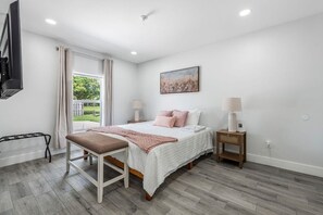 Ein Schlafzimmer mit einem Bett, Nachttischen, einer Bank, einem Fernseher und einem Fenster mit Vorhängen.