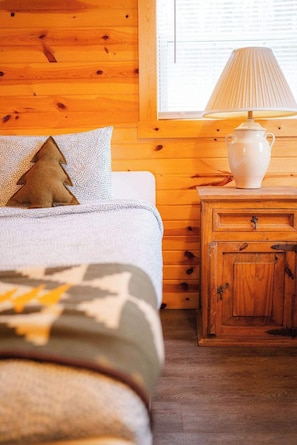Une chambre confortable avec une tête de lit en bois, un lit recouvert d’une couverture à motifs, une petite table de chevet en bois avec une lampe, et une fenêtre avec des stores.