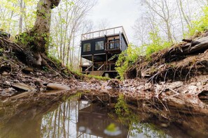 A cabin-like structure in a forested area with a reflective water body in front. - Unique Container Retreat With Creek Villa (McArthur)