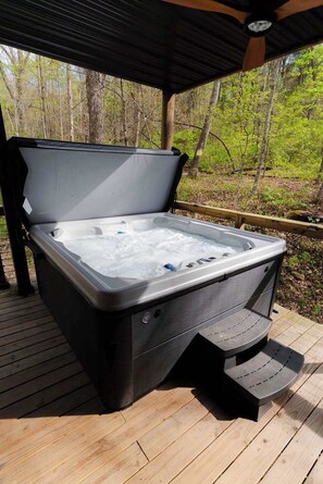 A hot tub on a wooden deck with a covered area overhead, surrounded by trees. - Unique Container Retreat With Creek Villa (McArthur)