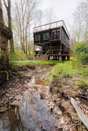 Una cabaña de varios niveles con un deck de madera y un pequeño arroyo en la parte delantera.