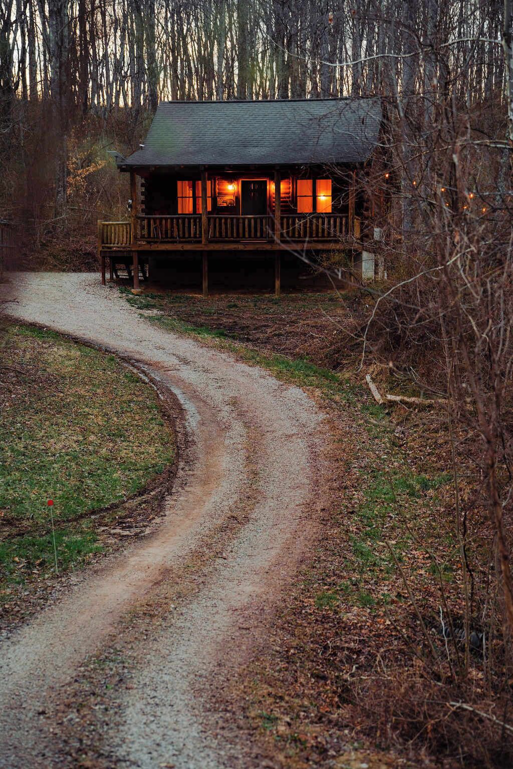 Una cabaña con un porche, un sendero de tierra que la lleva a ella, y un fondo boscoso.