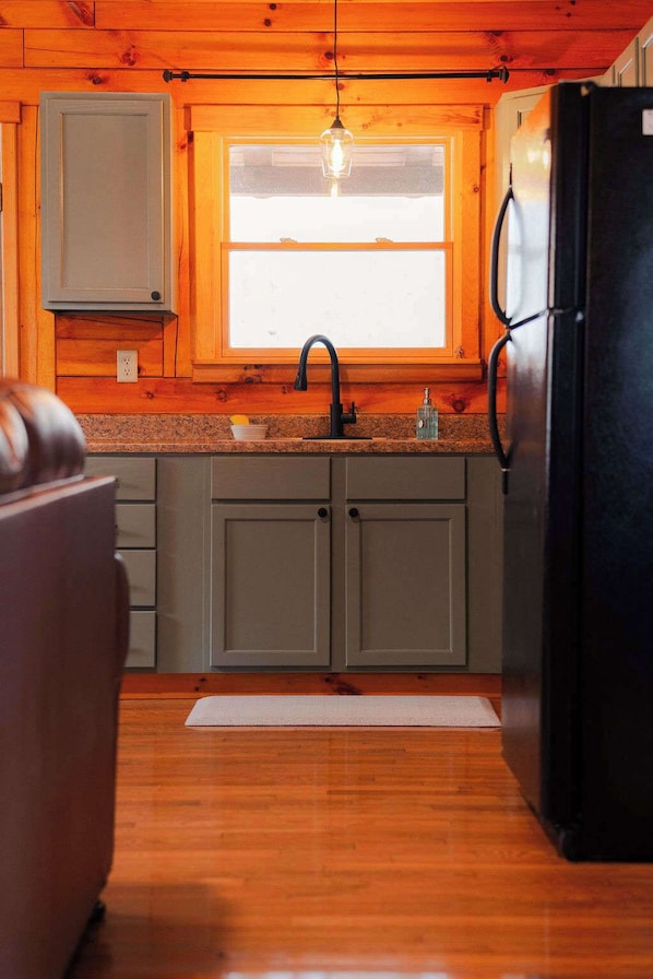 A kitchen with wooden cabinets, a black refrigerator, a window with a light fixture, and a countertop. - Secluded Cozy Log Cabin Villa (McArthur)