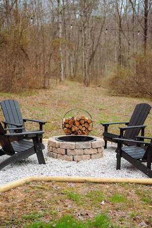 Two Adirondack chairs facing a fire pit with a log stack in the center, surrounded by a gravel area with a hose in the foreground. - Secluded Cozy Log Cabin Villa (McArthur)