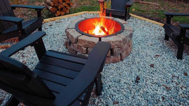 A fire pit surrounded by Adirondack chairs, set on a gravel surface with trees in the background. - Secluded Cozy Log Cabin Villa (McArthur)