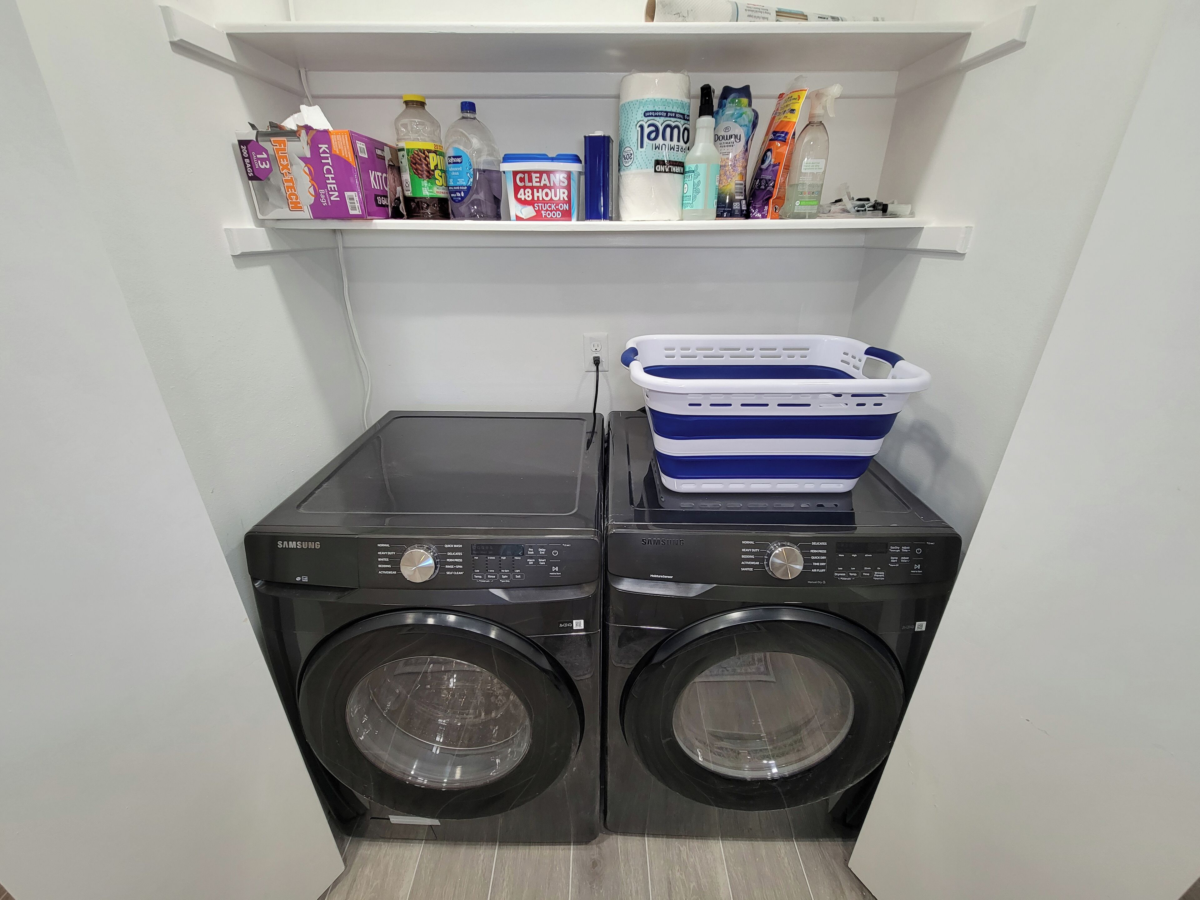 A laundry area with two stacked washing machines and a shelf above containing various cleaning supplies.