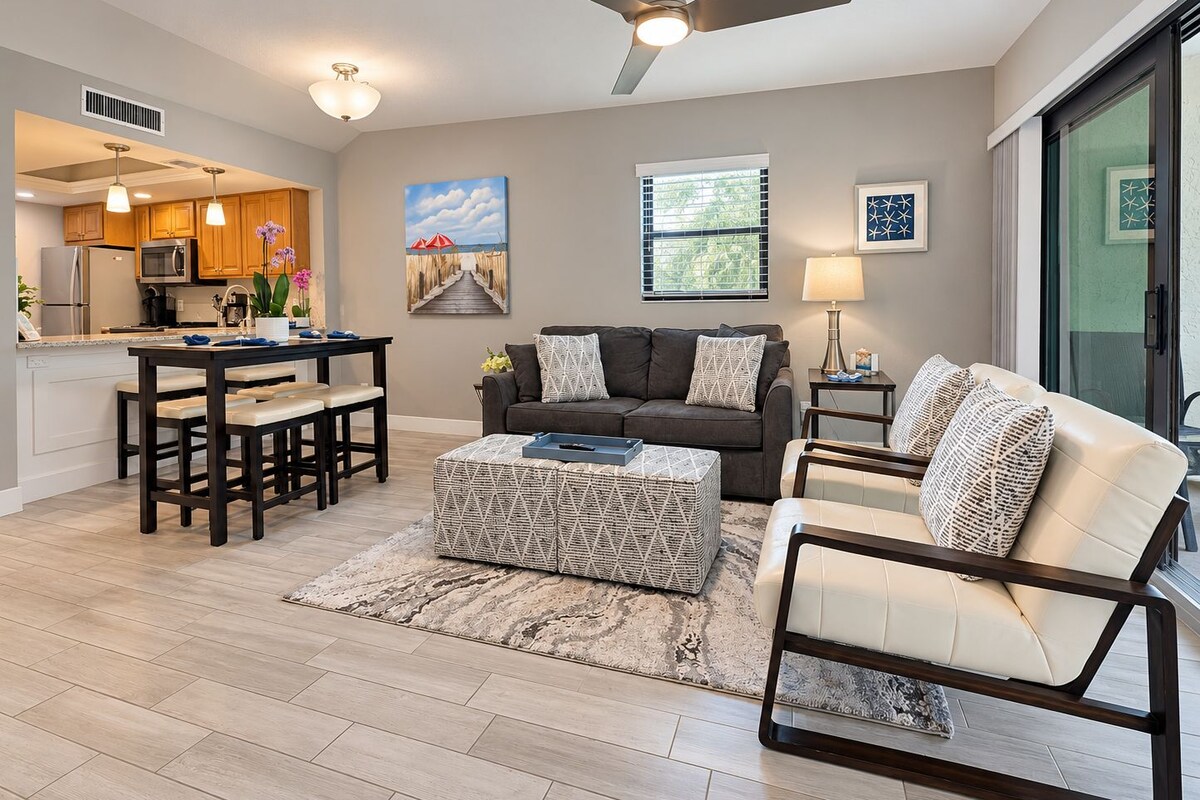 A modern living room with a sofa, ottoman, and coffee table, a dining area with a table and chairs, and a kitchen with appliances and cabinets.