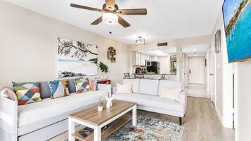 A modern living room with a white sofa, colorful cushions, a wooden coffee table, and a ceiling fan.