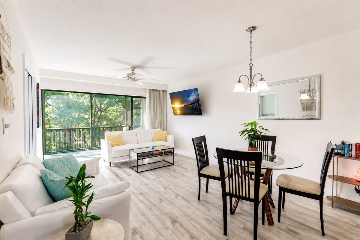 Sala moderna com sofá branco, mesa de jantar com cadeiras, mesa de centro e uma janela grande com vista para o verde.