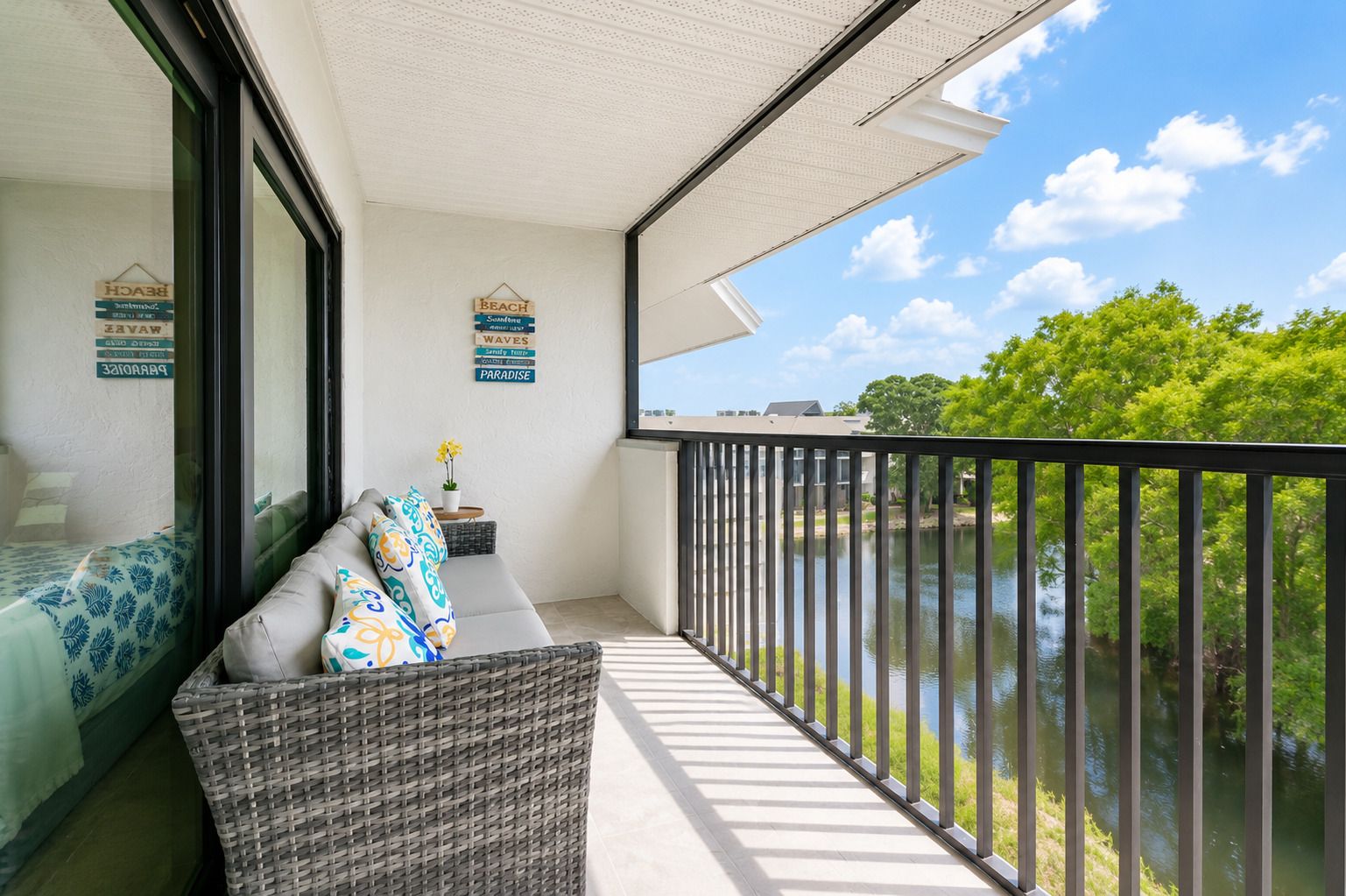 Un balcon donnant sur un plan d’eau, un canapé en osier agrémenté de coussins à motifs et une petite table sur laquelle est posée une plante en pot.