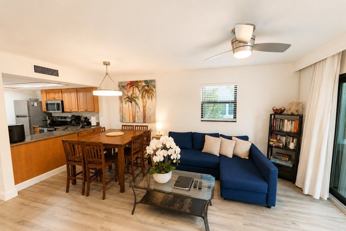 Uma sala de estar moderna com um sofá azul, uma mesa de jantar com cadeiras, uma mesa de centro, uma cozinha com armários de madeira e um ventilador de teto.