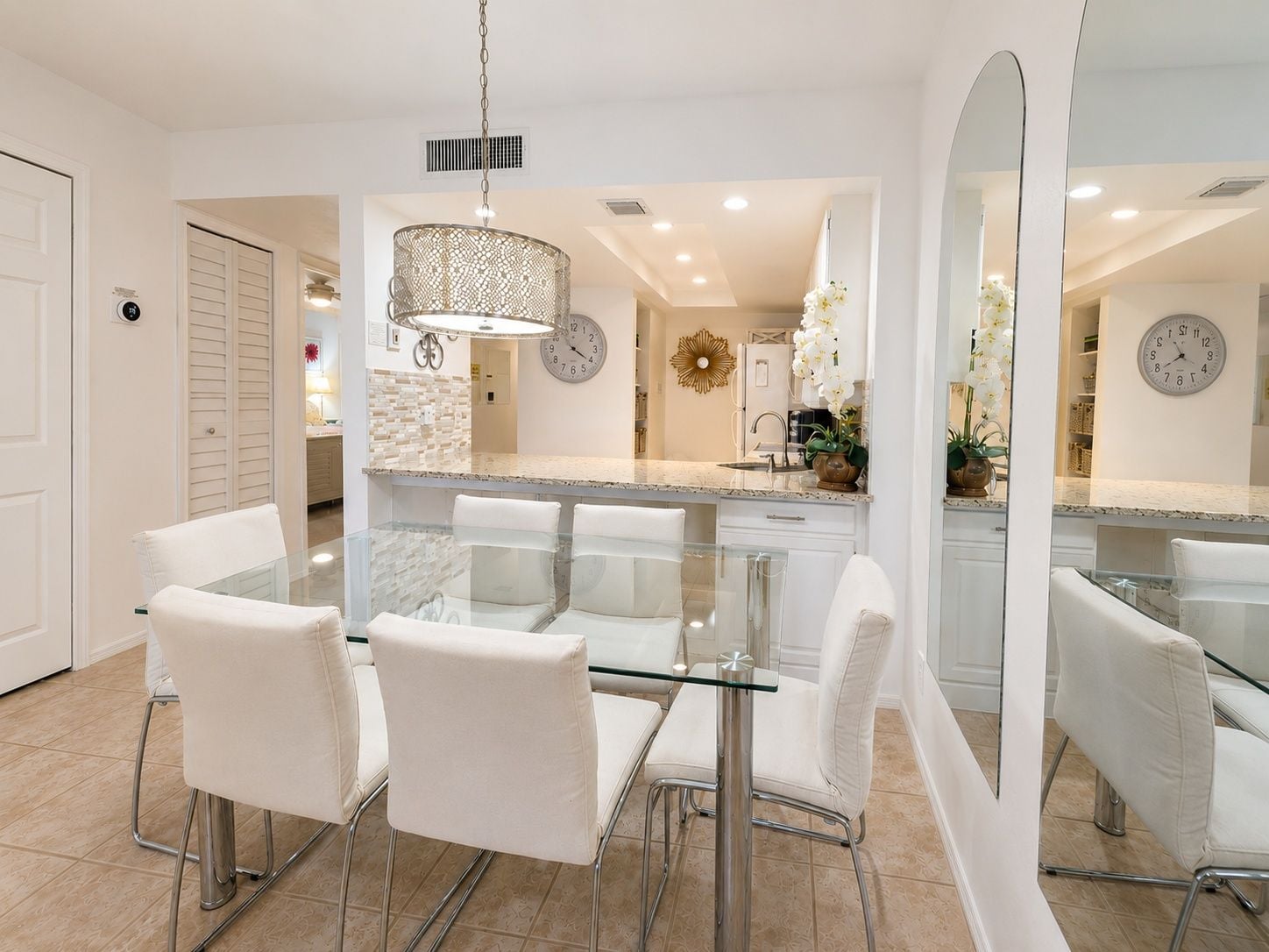 A modern kitchen with a glass dining table, white chairs, and a large mirror.