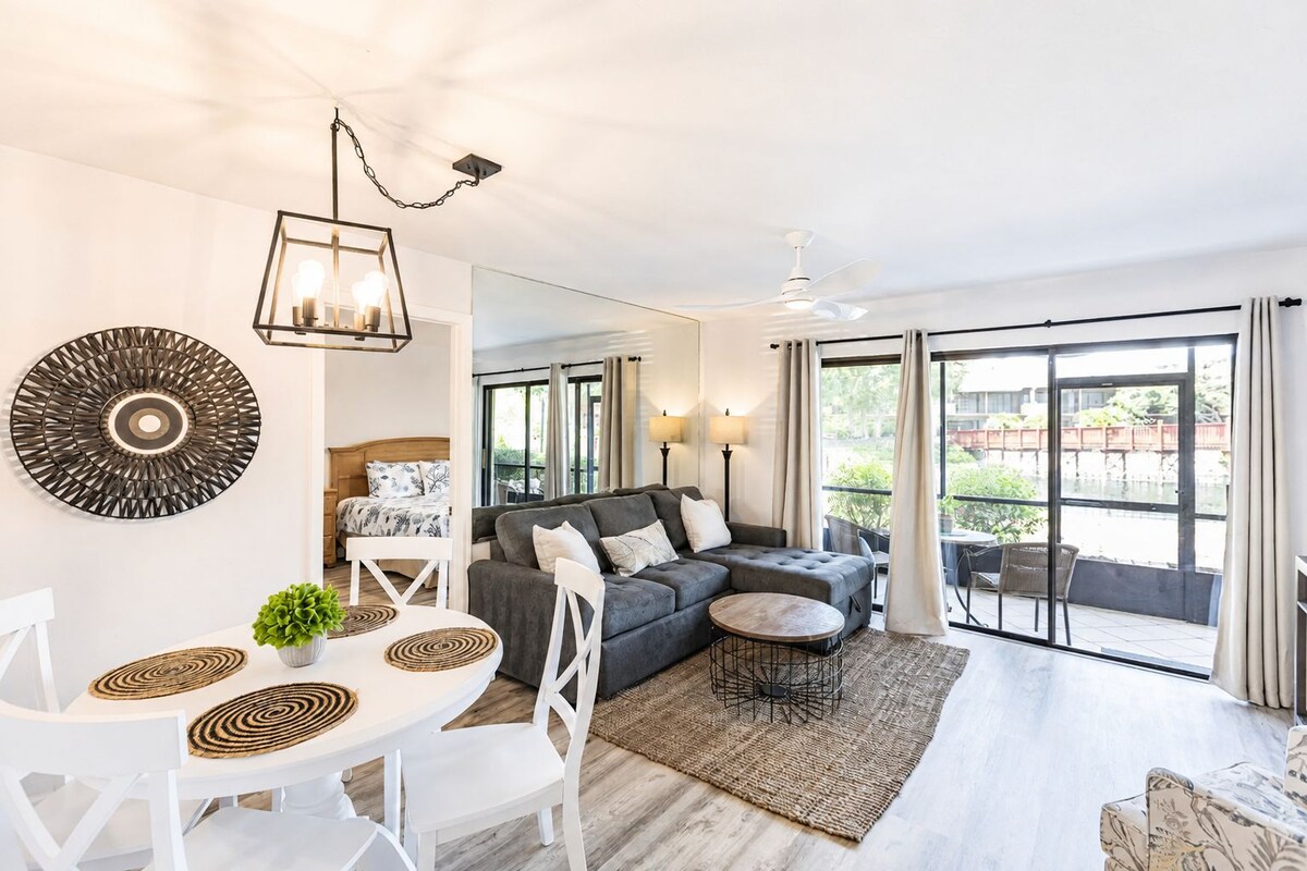 Sala de estar moderna com sofá, mesa de jantar, cadeiras, ventilador de teto e grandes janelas com vista para o exterior.