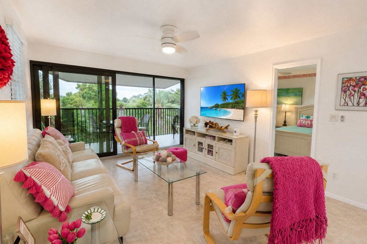 Uma sala de estar moderna com sofá, cadeiras, mesa de centro e televisão. Há uma varanda com vista para vegetação e uma praia.