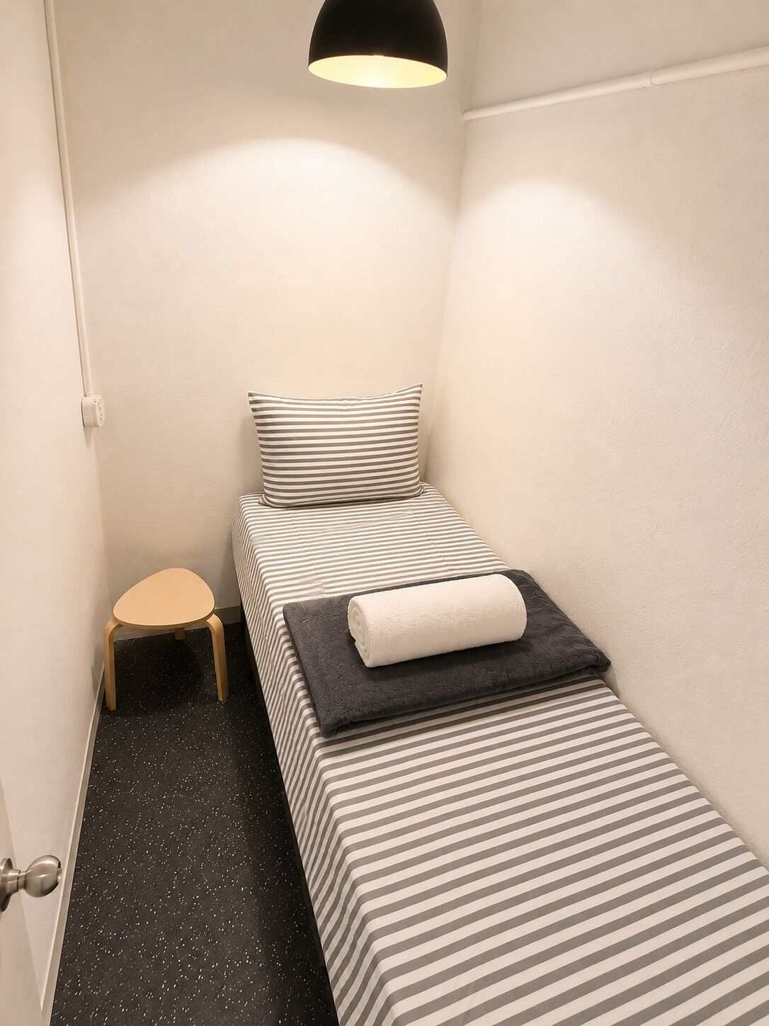 Une petite chambre avec un lit simple, une couette à rayures, un petit tabouret et une lampe noire.