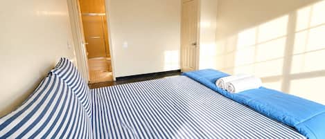 Quarto de hotel com uma cama, um travesseiro listrado em azul e branco, um cobre-leito azul e uma toalha branca enrolada.
