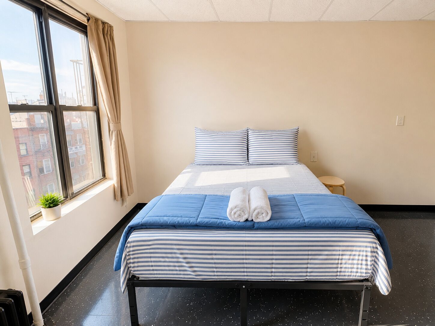 A hotel room with a bed, a small bedside table, a potted plant, and a window with curtains.