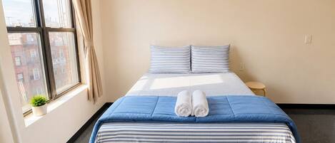 A hotel room with a bed, a small bedside table, a potted plant, and a window with curtains.