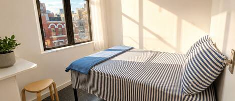 Een slaapkamer met een bed, een klein bureau, een stoel, een kamerplant en een raam met uitzicht op de stad.