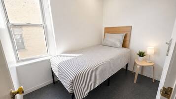 A small, single-bed room with a striped bedspread, a wooden headboard, a bedside table with a lamp, and a window with a view of a building.