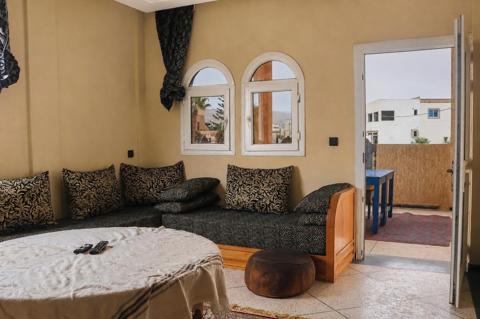 A cozy room with a sofa, a round table, and a wooden bench. There are two windows with arched tops and a door leading outside.