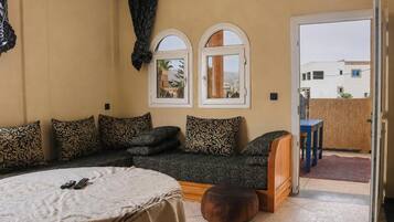 A cozy room with a sofa, a round table, and a wooden bench. There are two windows with arched tops and a door leading outside.